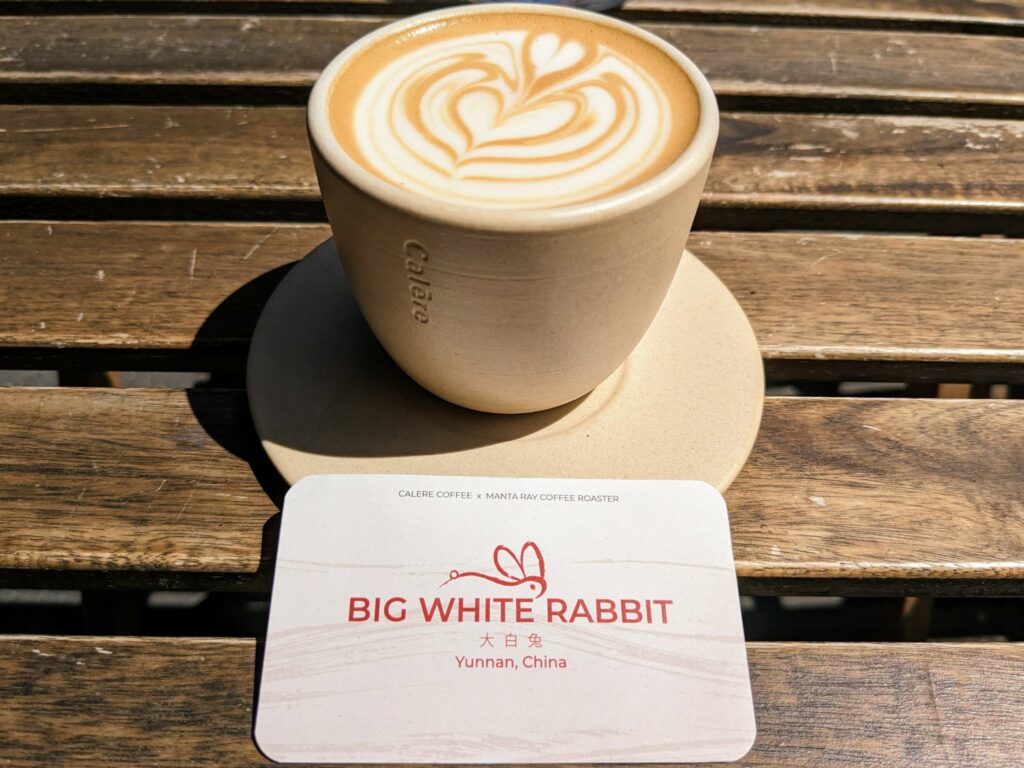 Full cup of coffee and saucer on wooden table, with a small card in front that says "Big White Rabbit, Yunnan, China"