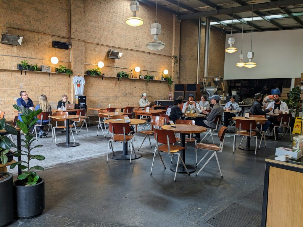 Interior of Everyday Coffee cafe, with several tables and chairs with people talking and working on laptops at front, and commercial coffee roasting equipment at rear