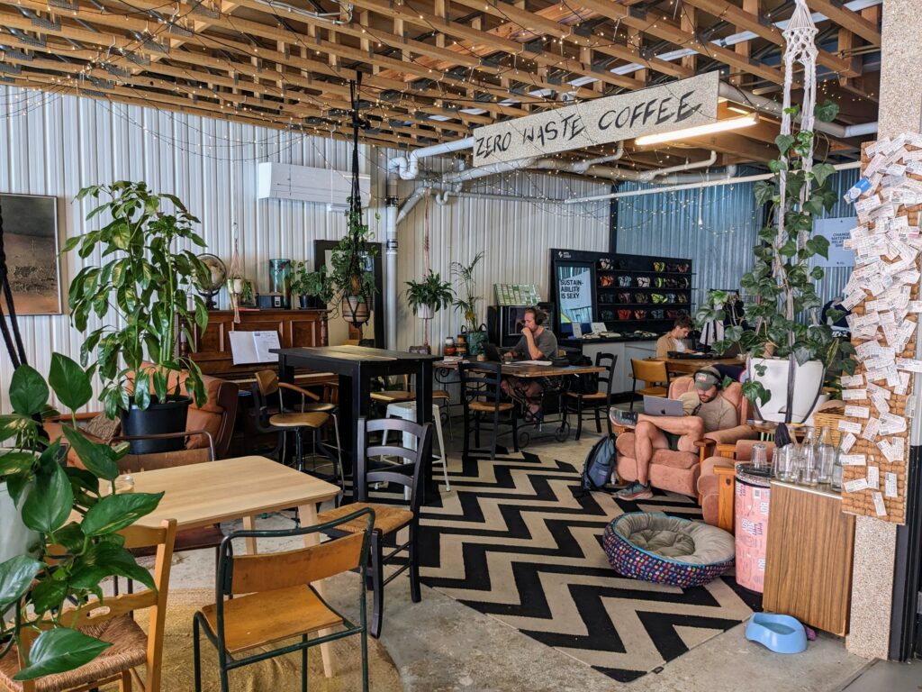 Interior view of Into Coffee, with a large sign below the rafters saying "Zero Waste Coffee". Several tables and chairs, a piano, potted plant, and people working on their laptops.