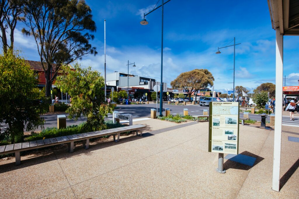 Street scene in Sorrento, Mornington Peninsula, with shops and cars in the background and an information board about historical Sorrento on the footpath in the foreground.