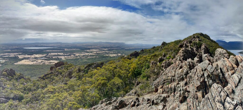 Panoramic view from the top of Boronia Peak near Halls Gap in the Grampians, with rocky ridgeline in foreground, tree-lined mountain slopes in middle distance, and lakes and farmland in far distance