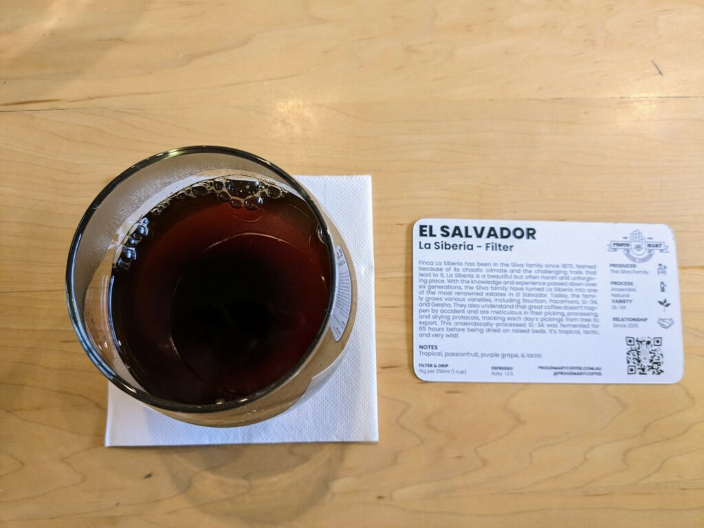 Overhead view of a glass of black coffee on a wooden tabletop, with a card of tasting notes alongside that's headed up with "El Salvador La Siberia - Filter"