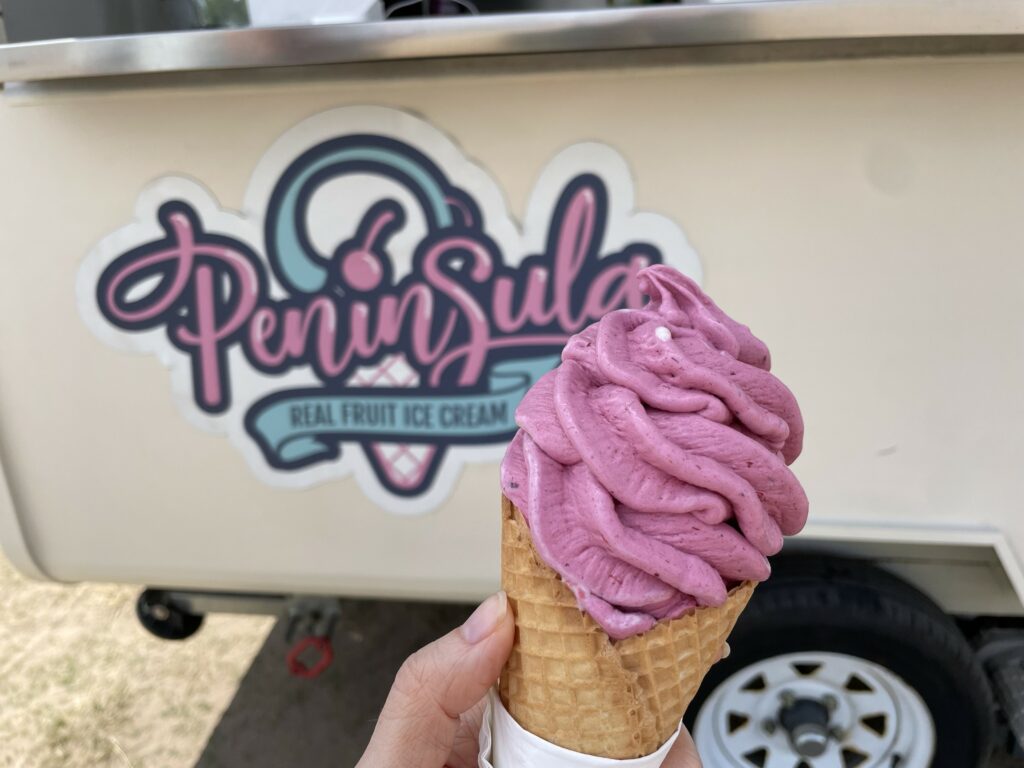 Ice cream in a cone being held in hand in front of a caravan with "Peninsula real fruit ice cream" on the side