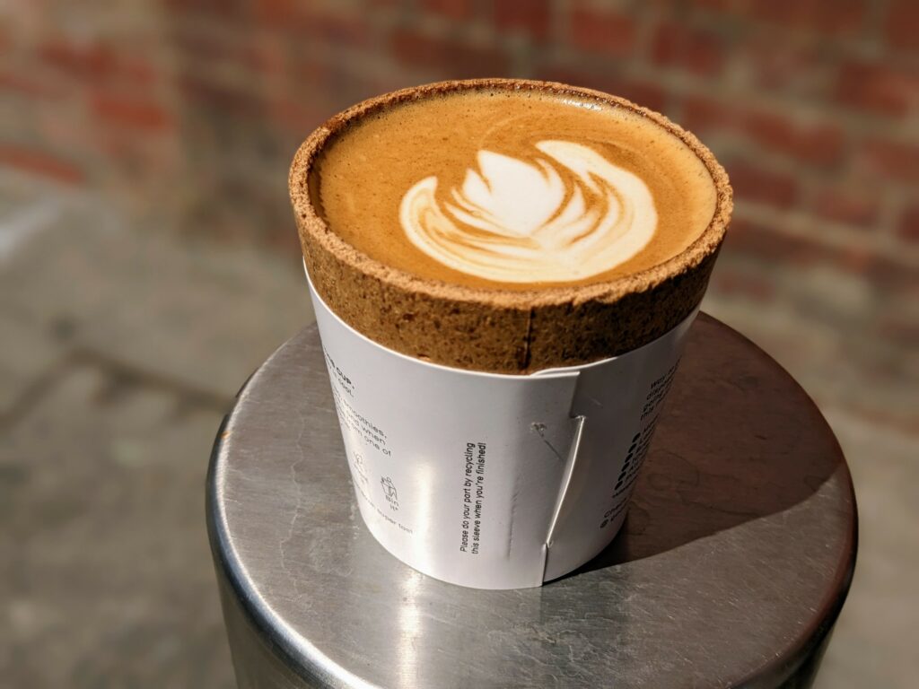 Coffee with latte art in edible cup, sitting on a metal bollard with a blurred background of bricks and stone tiles