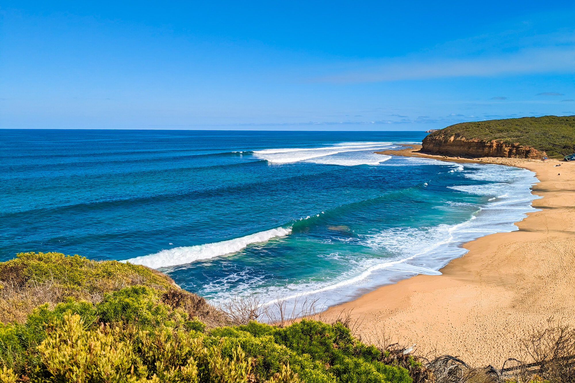 Bells Beach from the viewing platform on a cloudless, sunny day. The perfectly-formed waves are showcasing why this beach is popular with surfers, while the untouched sand indicates it's too windy for sunbathing