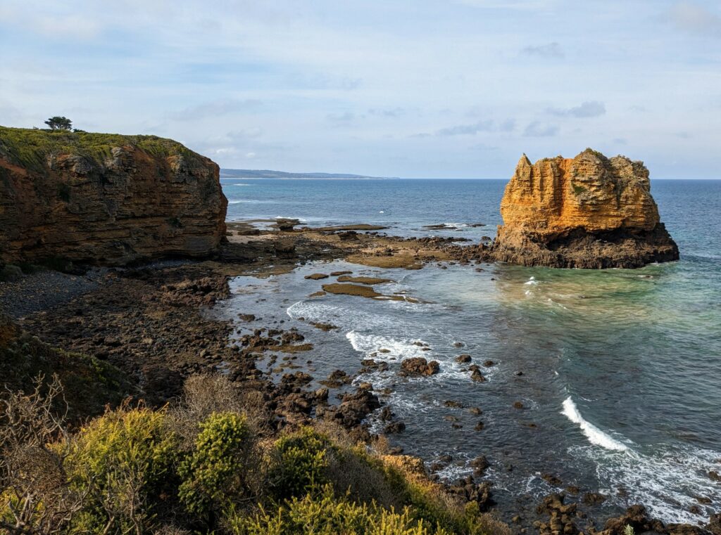 Large limestone rock sitting in the ocean just offshore, with rocky beach and small limestone cliff on the shore. Bushes in the foreground and distant land on the horizon