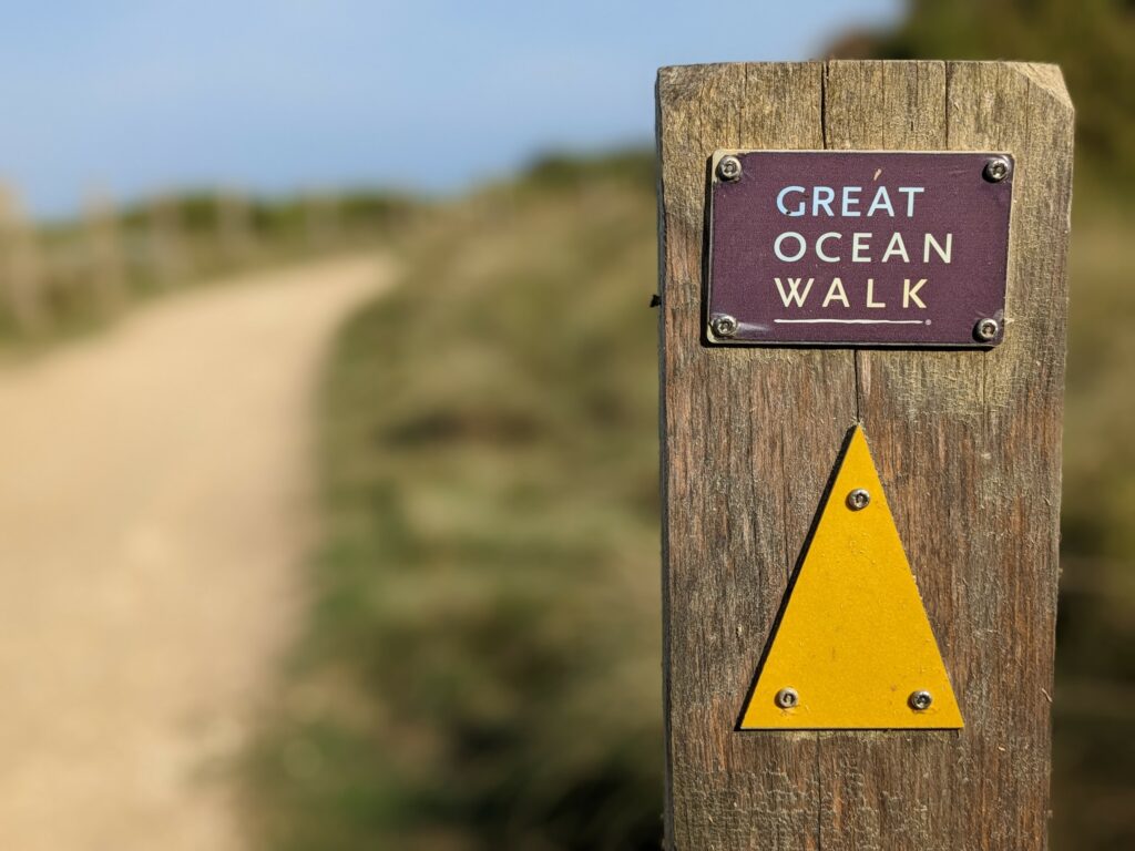 Wooden trail marker for the Great Ocean Walk, with a yellow triangle pointing the way. Blurred path in the background
