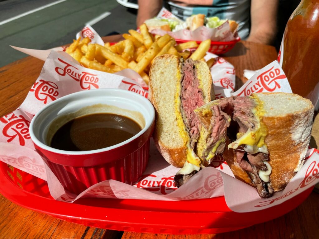 French dip roast beef sandwich sitting in red plastic tray on wooden outdoor table, with bowl of fries in the background