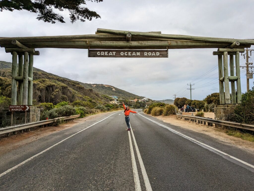 Young woman in an orange jacket standing in the road under a large stone and wood memorial archway that spans the Great Ocean Road. A small carpark is visible to the right beind the arch. A small sign saying "Historical Marker" is visible on the left hand side of the arch, and a larger sign saying "Great Ocean Road" hangs from the top of the arch.