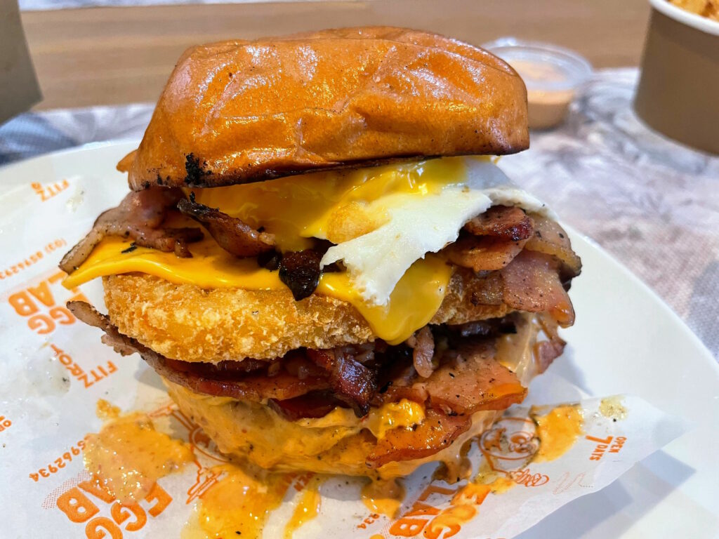 Large sandwich on a plate - two burger buns with egg, bacon, cheese, and hash brown visible between them