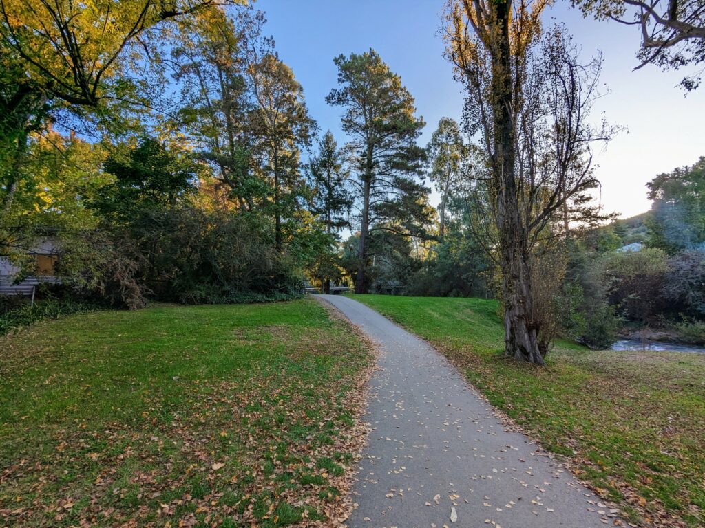 Paved path close to river, with a grassy lawn and autumnal trees on either side.