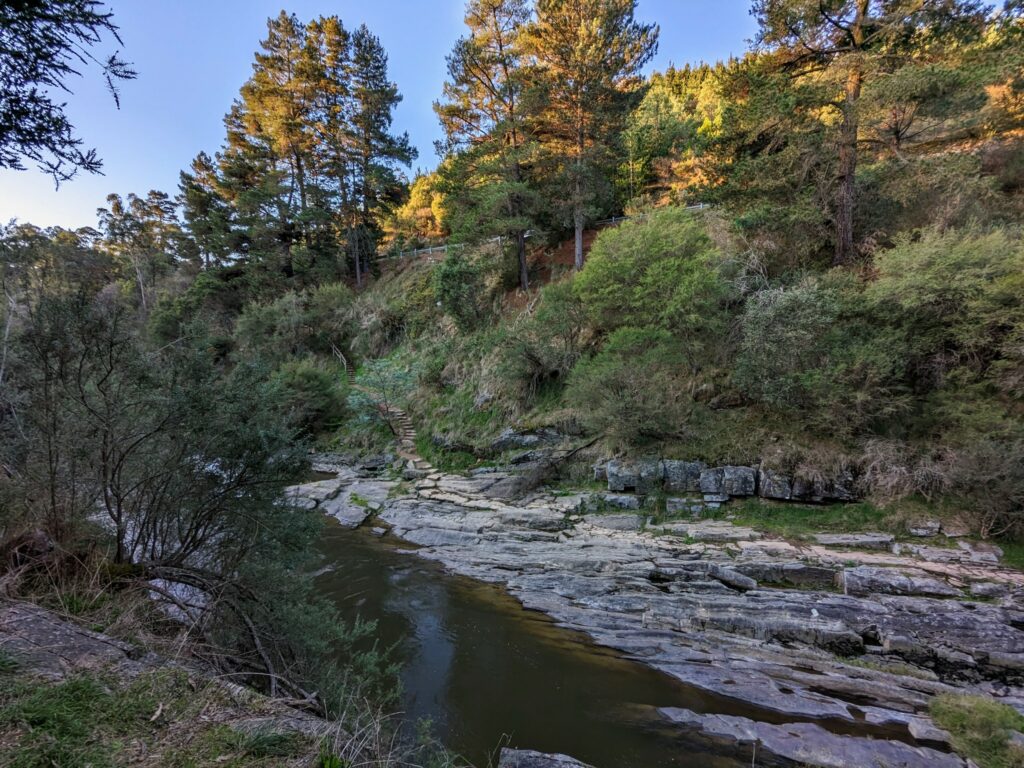 View over slowly-flowing river with large, flat rocks on opposite bank. Path leads down a staircase in the background to cross the rocks