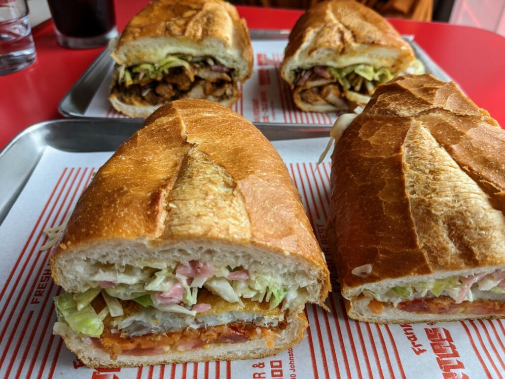 Torta-style sandwiches on trays on a red table: fish in foreground, al pastor in background