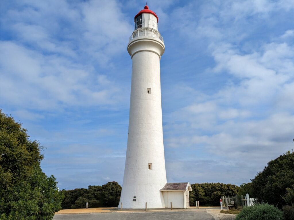 Tall white lighthouse in front of blue sky with a few scattered clouds, surrounded by low trees and bushes 
