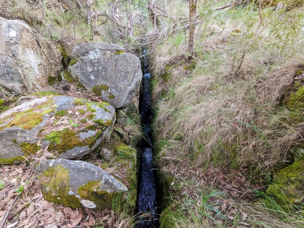 Tail race (narrow channel with water running through it) carved into rocks, with rocks and grasses on either side