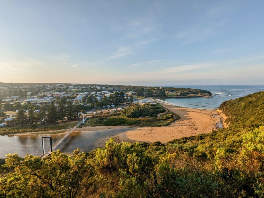 View from Port Campbell Lookout over beach, town, ocean, and suspension bridge, with trees in the foreground