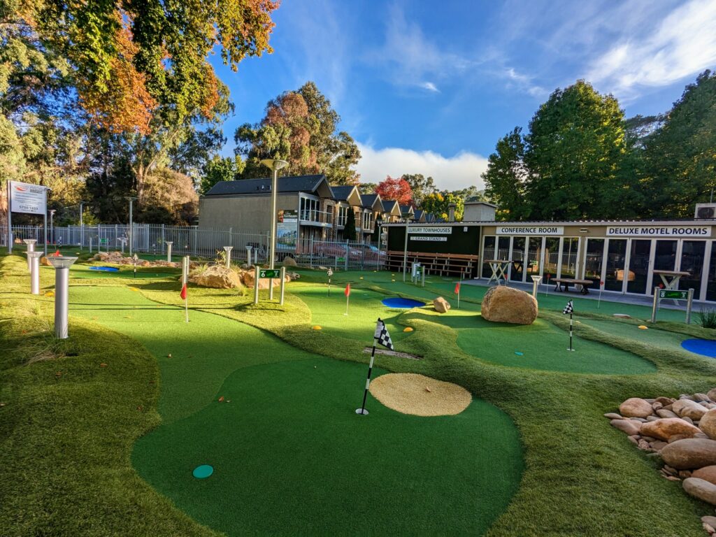 Mini golf course with several different holes visible. Holes marked by checkered flags. Townhouses and conference room visible behind the course.