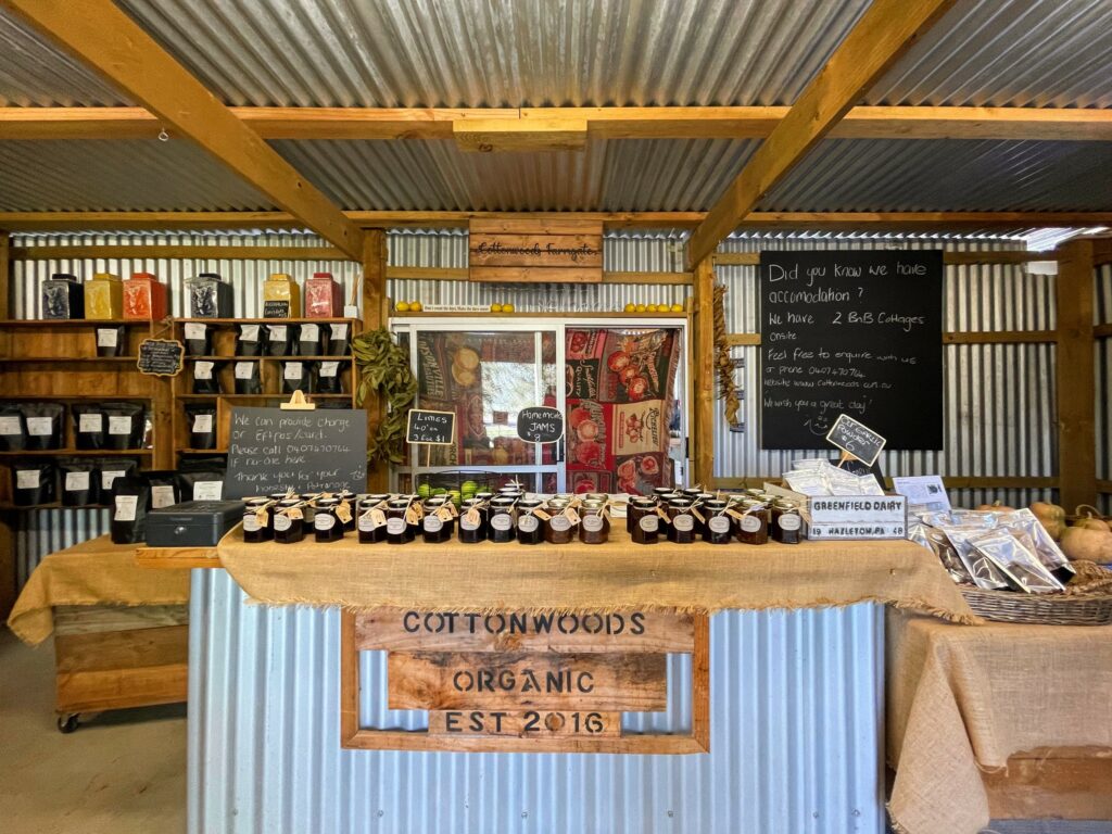 Interior of a small wood and corrugated iron farm shop, with sign saying Cottonwoods Organic Est 2016 on the counter. Jars of preserves on counter, with bags of dried fruit, pumpkins, limes, and other food items in background.
