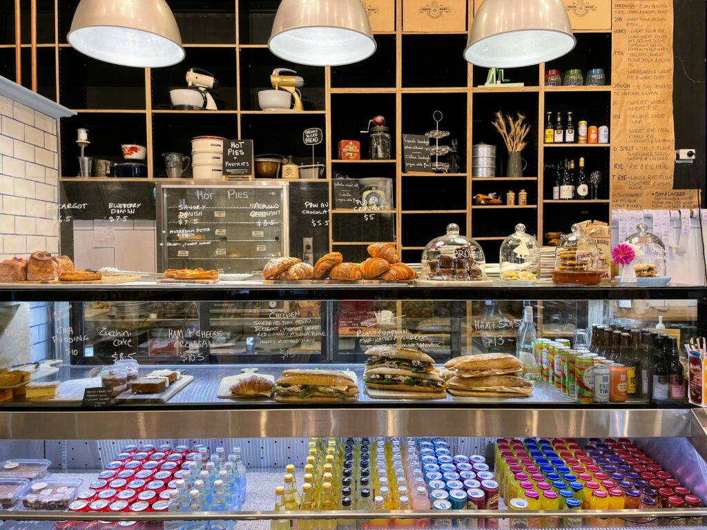 Food counter with baguettes, pastries, biscuits, and drinks for sale. Pie warmer in the background, along with shelves holding a range of items from retro cake makers to plants and bottles.
