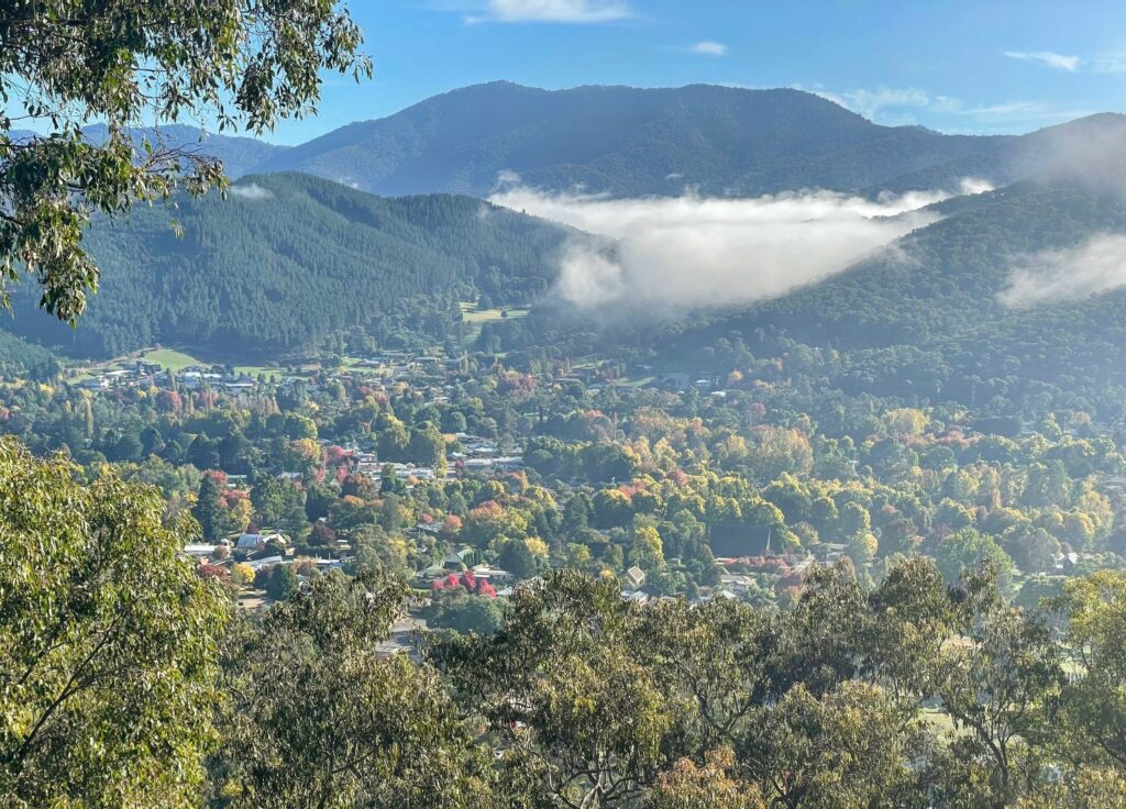 View from a lookout over a township and trees on a valley floor, with hills and mountains in the background. Blue skies with some fog visible in the valley