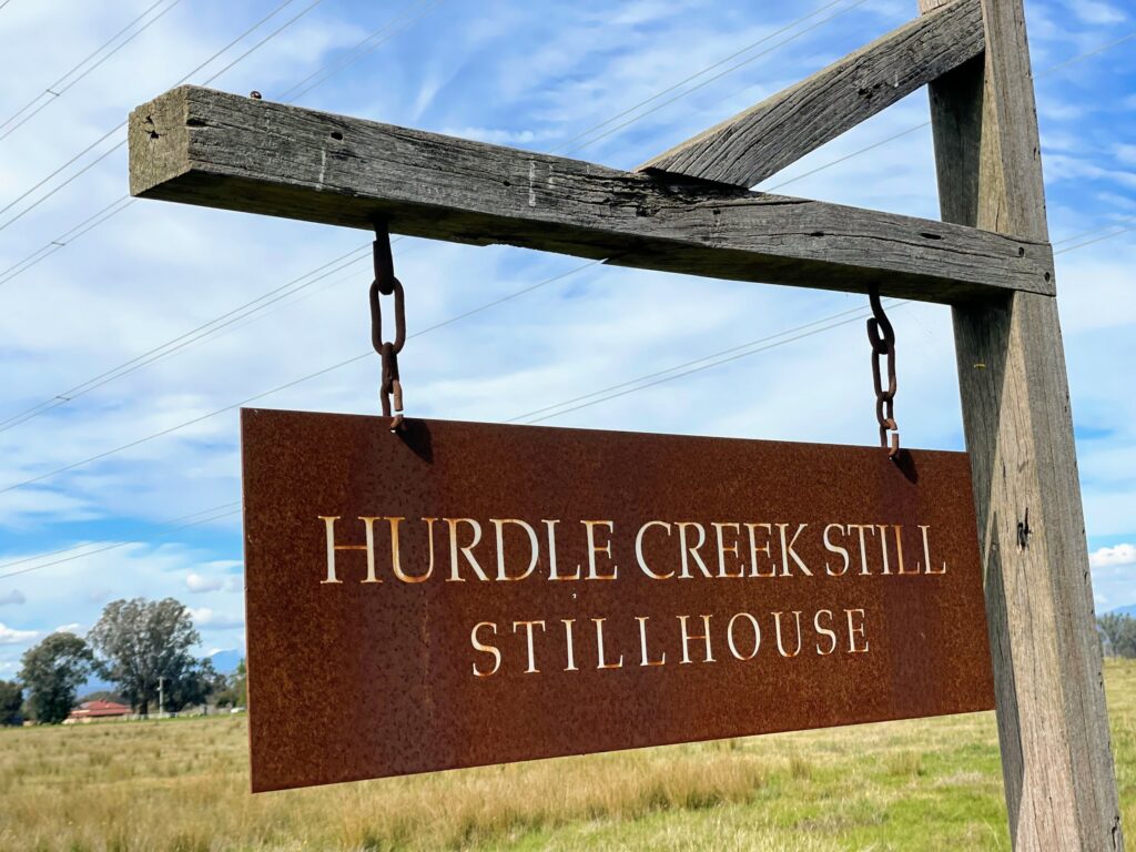 Rusty metal sign saying "Hurdle Creek Still Stillhouse", hanging from metal chains on wooden pole. Grassed paddock and trees in the background against blue sky.