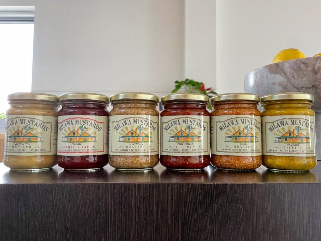 Jars of mustard, chutney, pickles, and chilli jam from Milawa Mustards lined up alongside each other on a wood-patterned bench
