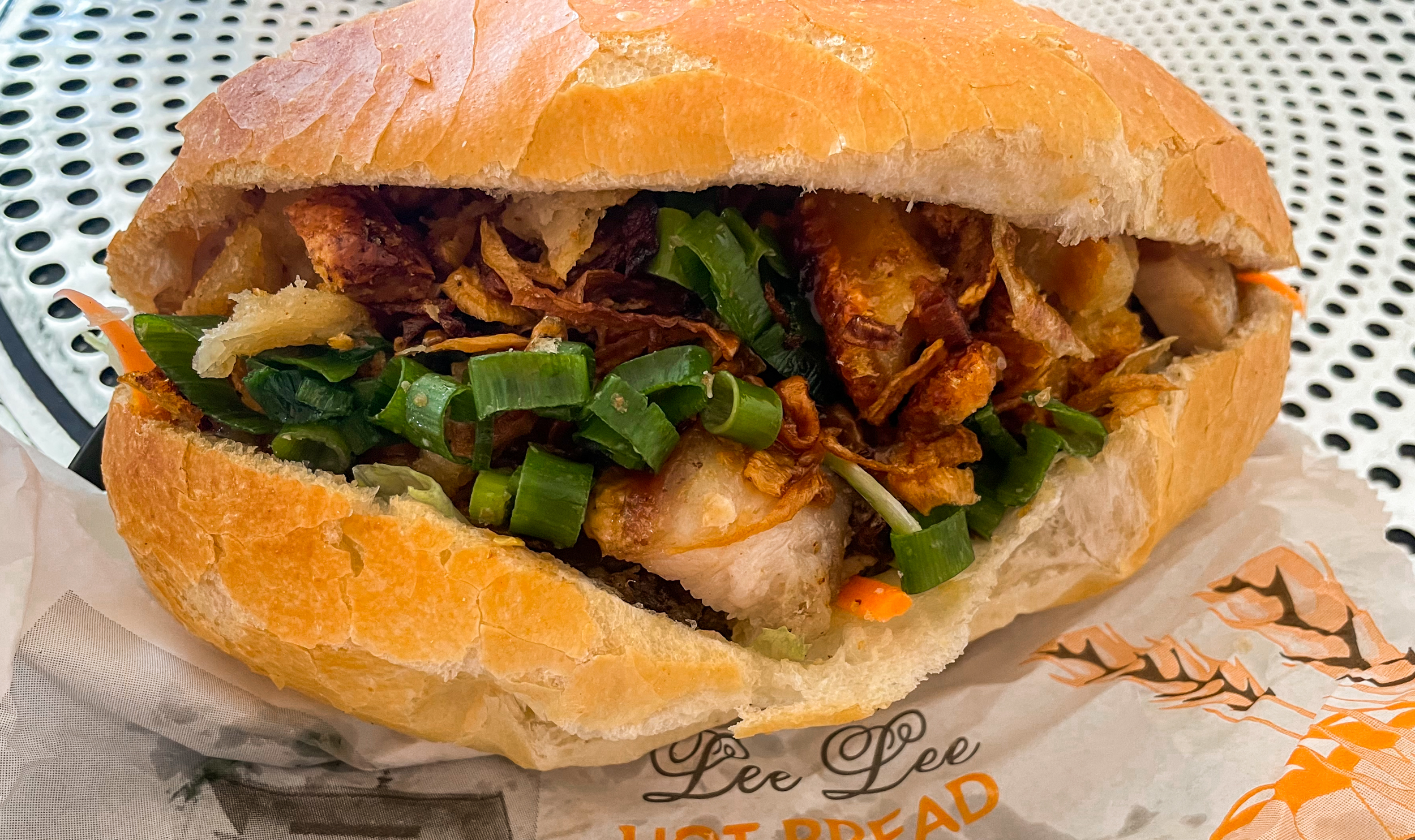Banh mi bread roll with pork, spring onion, and other fillings inside. Sitting on paper bag with Lee Lee Hot Bread printed on it