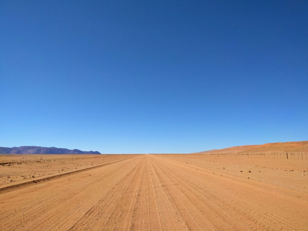 Long straight empty dirt road in Namibia, with small mountain range in distance.