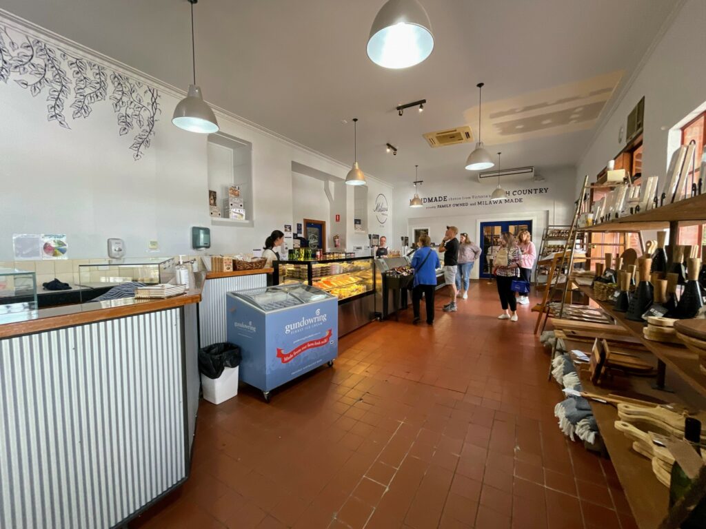 Inside of Milawa Cheese Company store, with cheese, deli meat, and icecream displays on left side and range of boards, bottles, and other items on right. Customers shopping at far end.