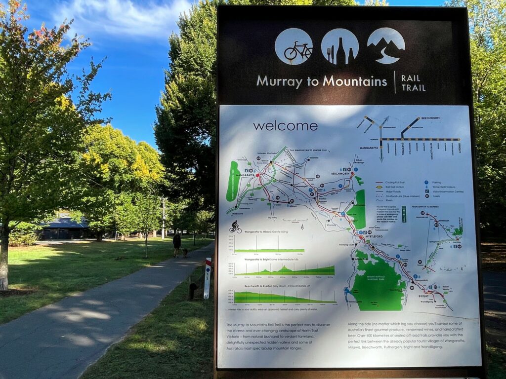 Large sign beside a paved path advertising the Murray to Mountains Rail Trail, with maps and information. Person walking dog on the path in the middle distance