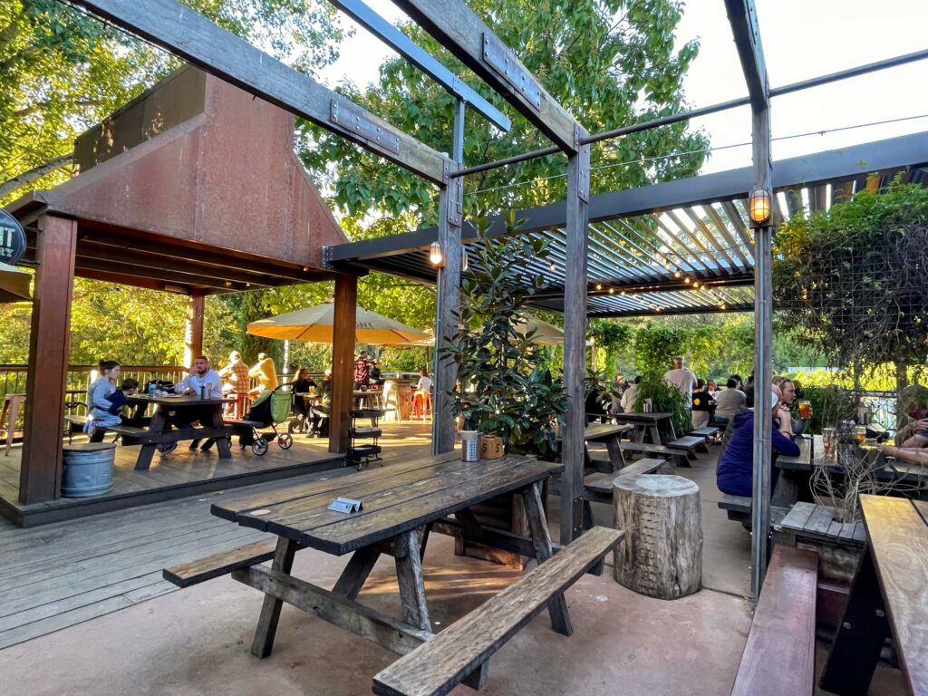 People sitting eating and drinking at outdoor tables on wooden and concrete decking outside a brewpub, with trees in the background
