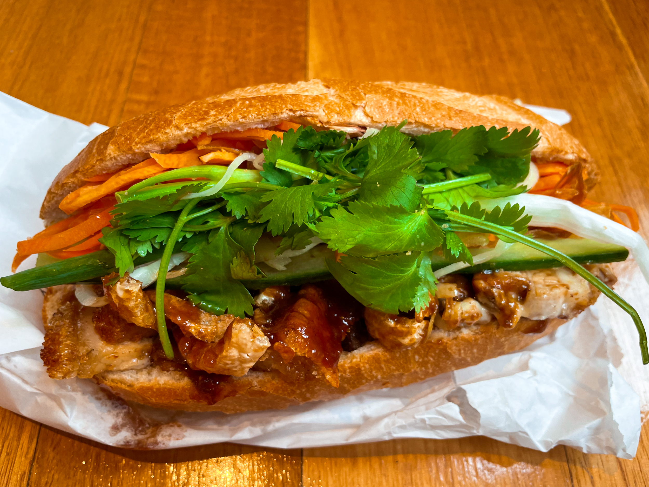 Banh mi bread roll with pork, coriander, and other fillings inside. Sitting on paper bag on wooden table