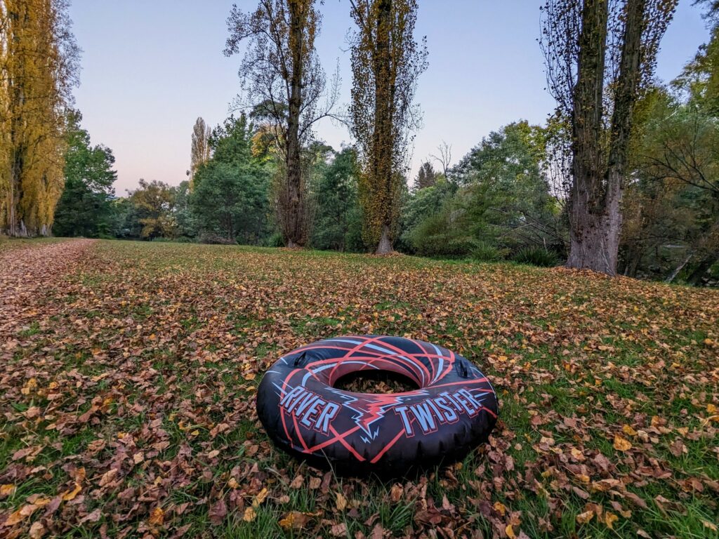 Inflatable ring with "River Twister" printed on it, sitting among autumn leaves near a river with a walking trail nearby and trees in the background.
