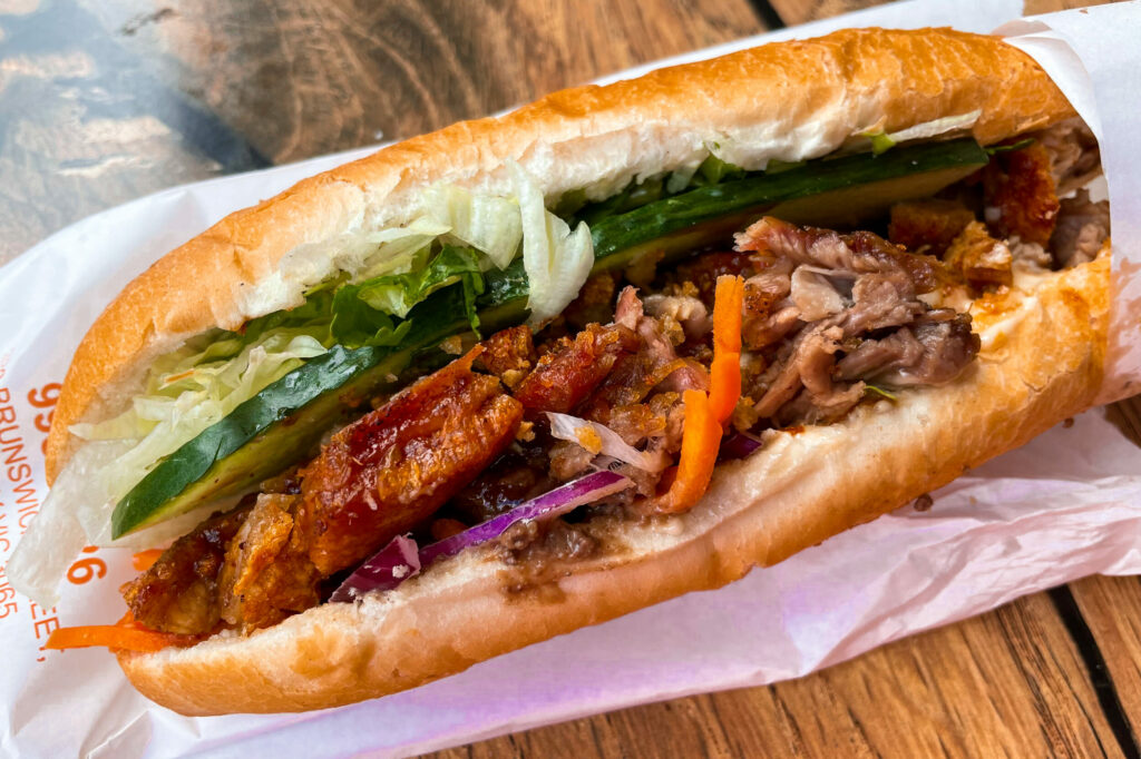A close-up of a crispy pork banh mi, laying atop a paper wrapper from Viet Rose in Fitzroy. Inside the fresh bread roll, layers of fried pork are squeezed up against slices of cucumber and lettuce