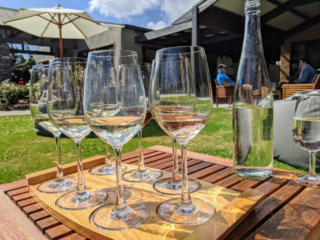 Seven partially-filled wine glasses on a wooden platter, sitting on a wooden table on the grass outside a winery. Beanbags and people in the background.
