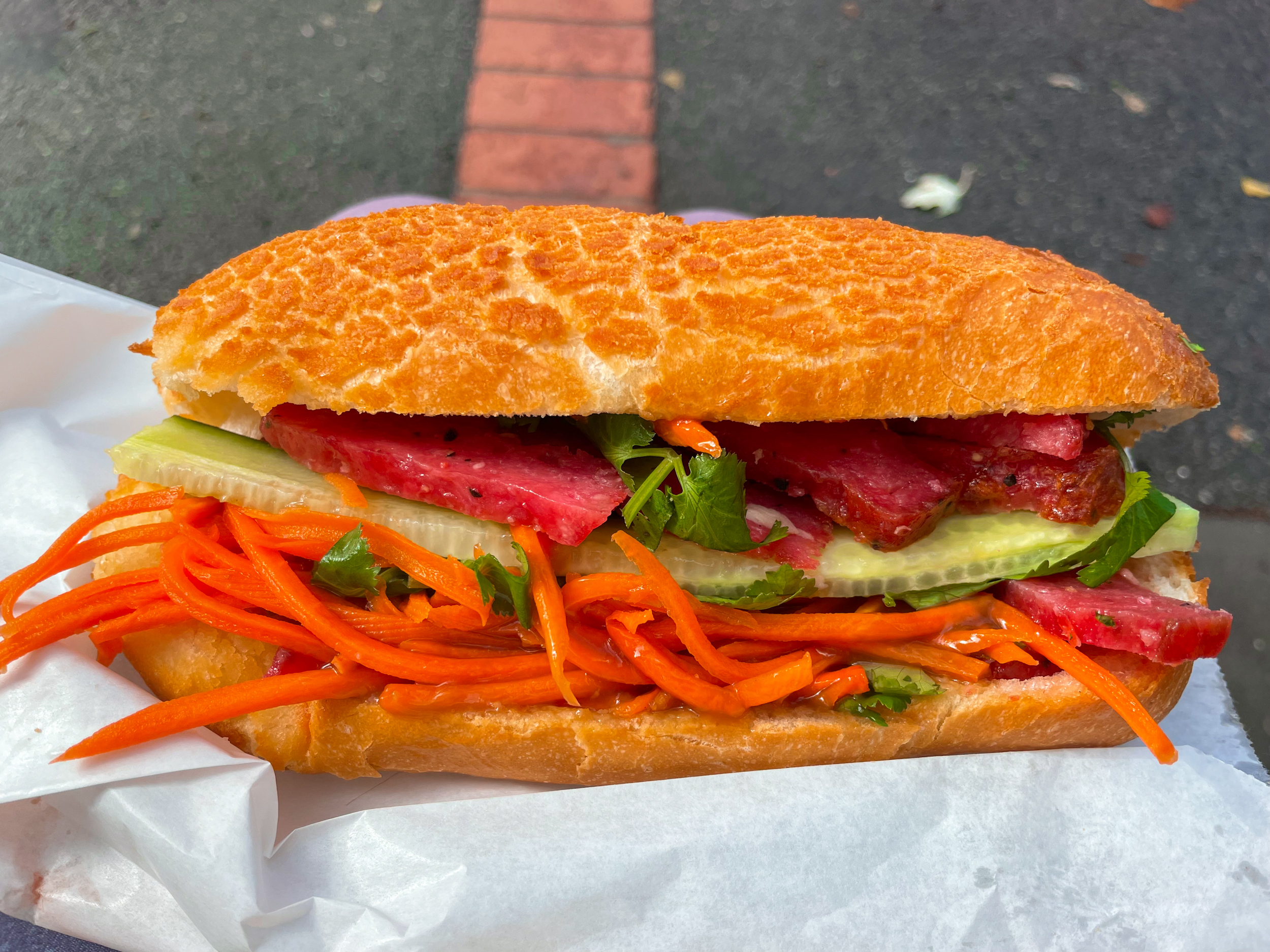 Banh mi tiger bread roll with pink pork, cucumber, and other fillings inside. Sitting on paper bag