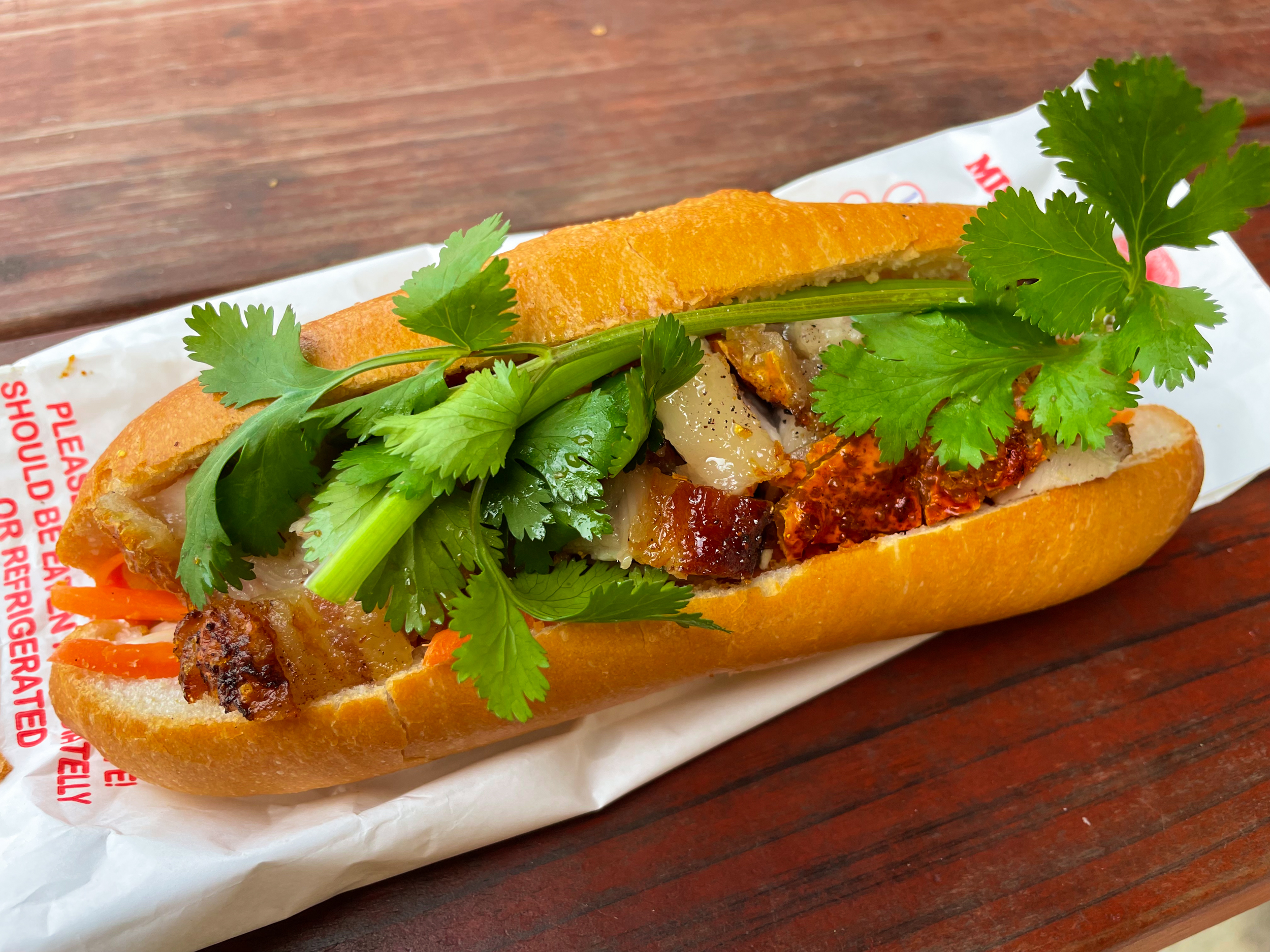 Banh mi bread roll with pork, coriander, and other fillings inside. Sitting on paper bag on table