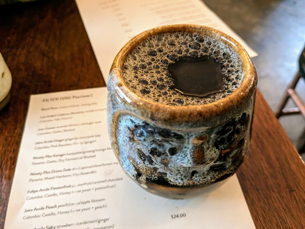 Black coffee in a small, decorated ceramic mug, sitting on a paper menu of pourover coffee varieties on a wooden table.