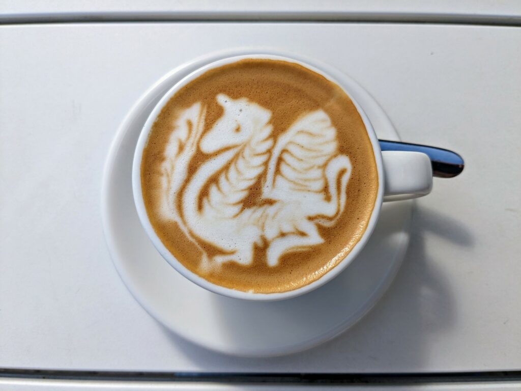 Overhead view of white cup with coffee and intricate latte art, on a white sauced and white painted wooden table.