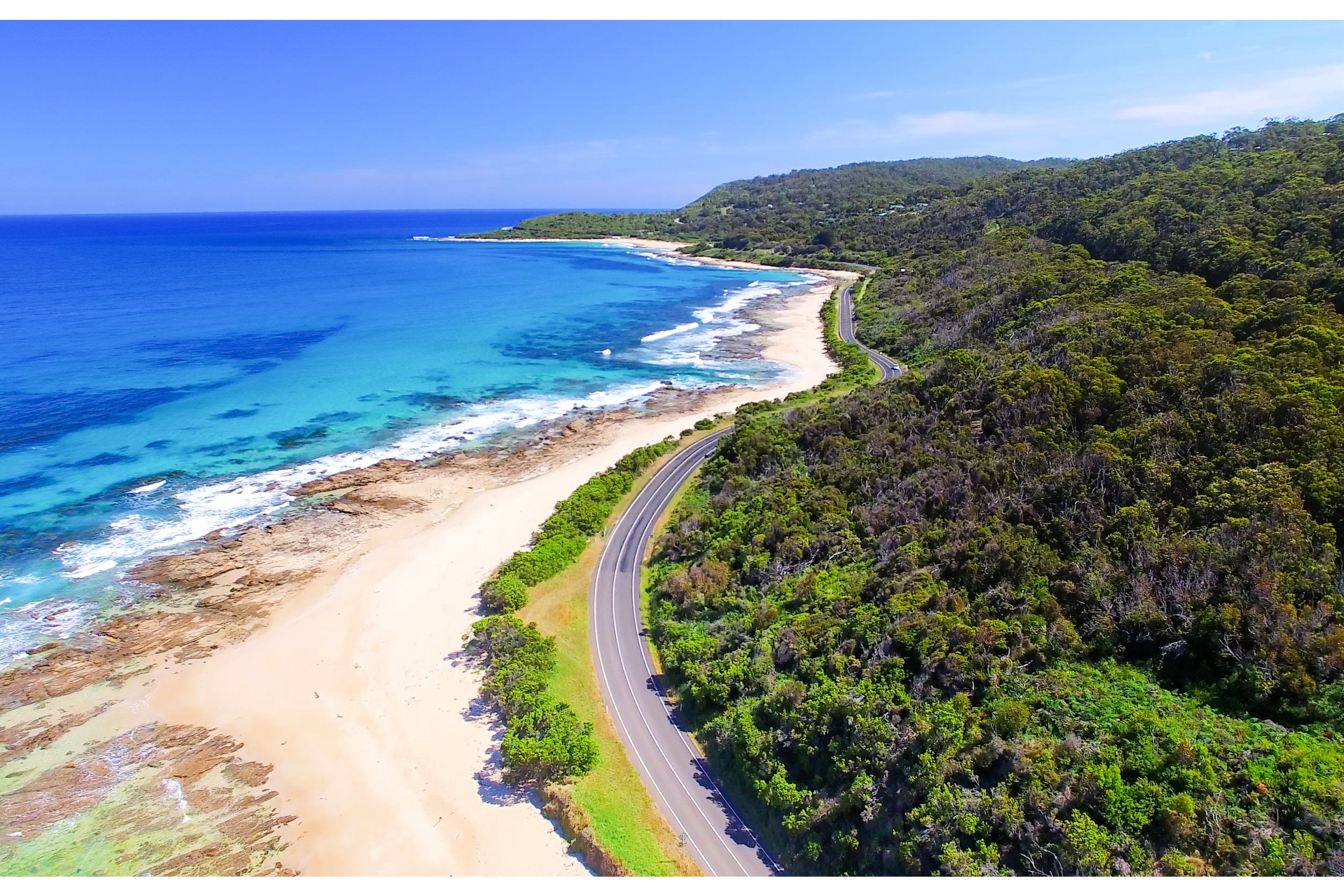Looking down on a coastal winding road on a perfect blue-sky day. To the left of the road, pristine white sand stretches into calm, turquoise waters. On the right, dense forest lines the hillsides