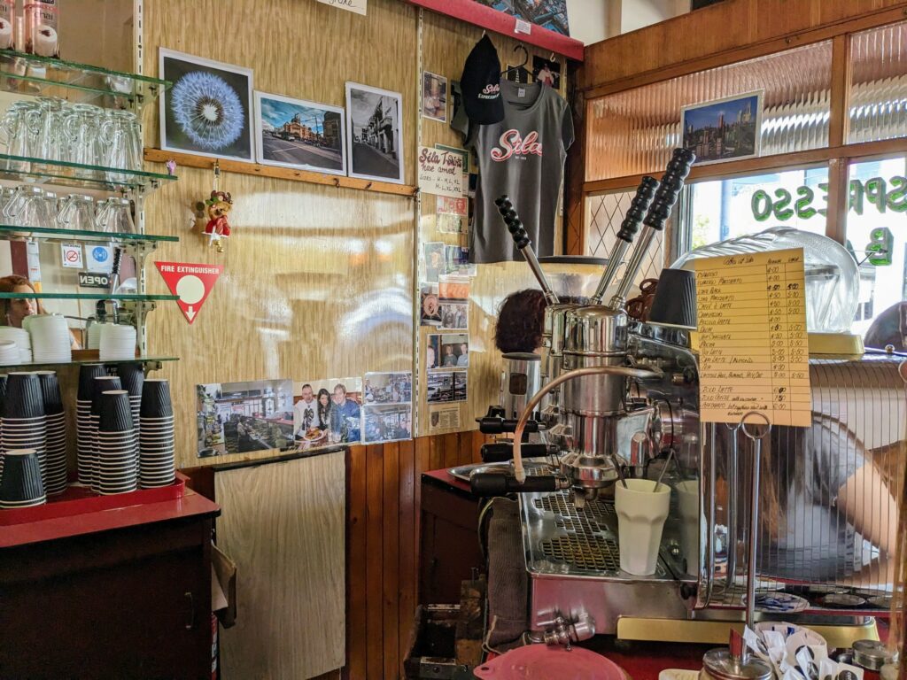 Counter area of an Italian-style coffee shop, with old lever-action espresso machine, photos on the wall behind, and cups and glasses stacked up.