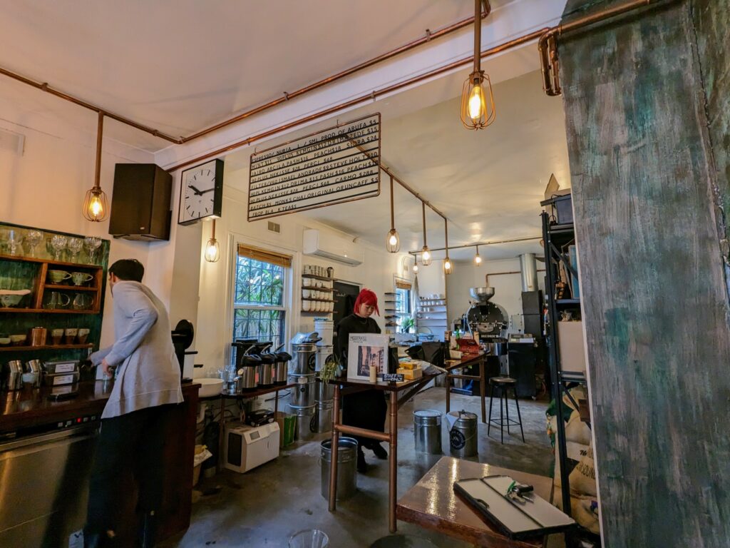 Interior of a small coffee roastery and cafe, with two staff members working. Industrial-style decore, with coffee roasting machine in the background, and large amounts of other coffee equipment on shelves and counters