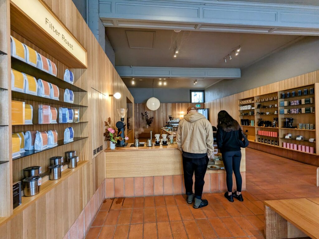 Interior of coffee shop with a warm, minimalist design. Wooden walls and furniture and paved floor. Coffee beans and equipment for sale on shelves on both sides, Counter in middle with range of coffee-making equipment. Two people standing at counter.