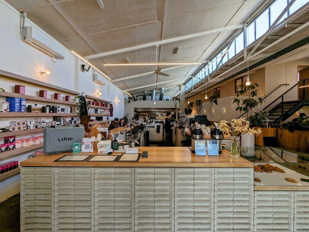 Interior of ONA Coffee, with large L-shaped counter and shelving running down the left-hand wall with coffee equipment for sale. Small glass display case on right with pastries.