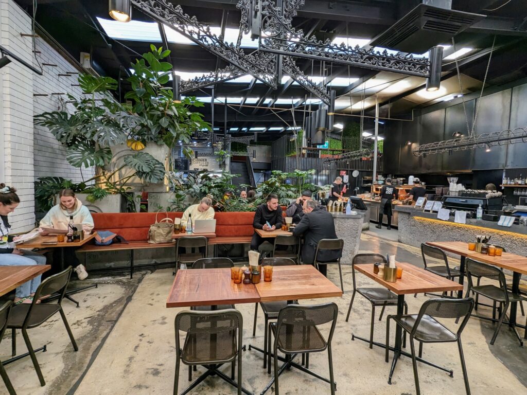Interior of Project 281 Coffee Roasters in Brunswick, Melbourne. Several tables and chairs in foreground, some occupied with customers. Large wrought-iron chandelier above. Many large plants in background. Counter to the right with staff making coffee and preparing food.