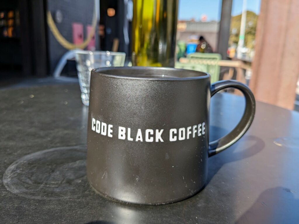 Black coffee mug with "Code Black Coffee" printed on the side, sitting on a black outdoor table. Water glass partially visible behind, with blurred people, table, and chairs in the background.