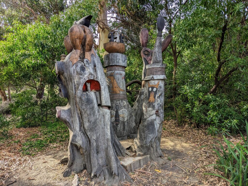 Carved and painted wooden sculpture of "Buckleys Bunyip", a mythical water creature. Set in natural surroundings with small trees and bushes nearby.