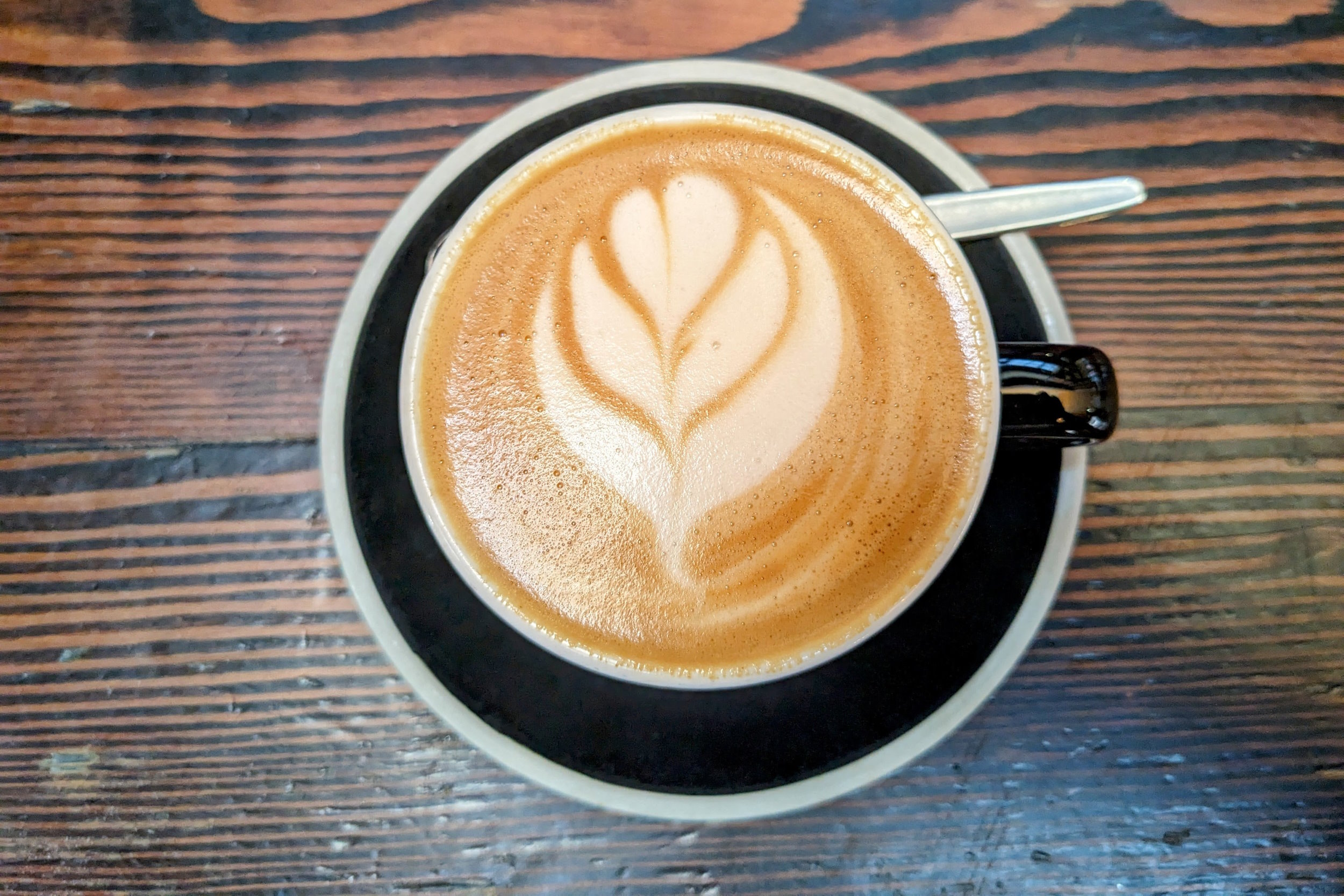 Overhead view of coffee with latte art, in a black cup on a matching saucer, sitting on a dark wood-grained table.