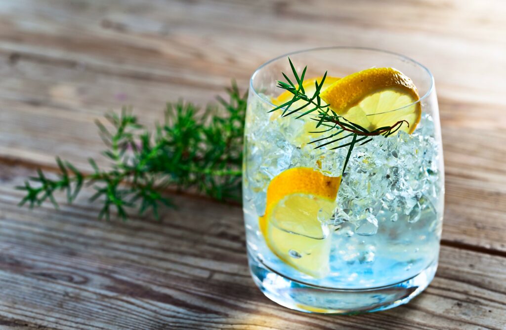 Small glass with gin, ice, lemon slices and juniper sprigs inside, on a wooden table