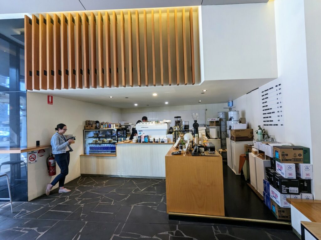 Inside of a small coffee shop with white walls and ceiling, and an L-shaped counter that's crowded with coffee equipment. A menu is visible on the right-hand wall, and one customer can be seen waiting for their order on the left.